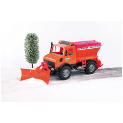 Orange Bruder MB-Unimog toy with snow plough clearing snow next to a snow-covered tree.