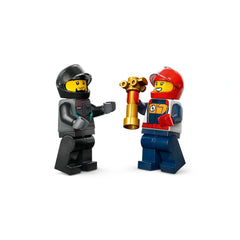 Two LEGO race driver minifigures celebrate victory, one holding a gold trophy. The first figure wears a black helmet and gray outfit, while the second dons a red helmet and colorful racing gear.