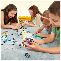 Four children sitting on a light grey carpet, engrossed in building with colorful LEGO bricks spread around them. One child holds a partially constructed vehicle, while others focus on their own creations. A bright yellow LEGO box is visible in the background, enhancing the playful atmosphere.