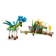 A whimsical green dream creature with flower decorations stands beside a colorful stable made of bricks, featuring a sloped roof, vibrant plants, and playful figures.