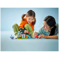 A young girl in an orange sweater and green pants joyfully plays with the LEGO® DUPLO® Mickey Mouse Clubhouse set, while an adult beside her interacts with a small red toy car on a blue background.
