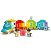 Colorful LEGO® DUPLO® Number Train set featuring a bright blue engine, numbered red and green cars, and two child figures holding a pink block with the number zero. A small white dog sits on the green car, creating an engaging scene for preschool learning.