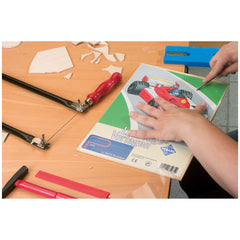 Hands working on a Prebaro Barnyard II fretsaw template, with red-handled fretsaw and cut wooden pieces on a table.