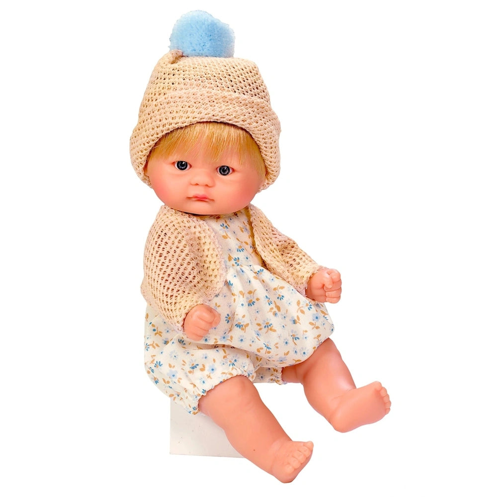 Asi Dolls Bomboncin doll in floral outfit, beige jacket, and hat with blue pom-pom sits on a white surface.