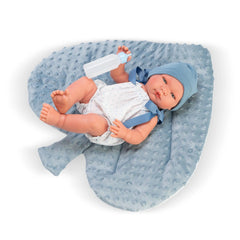 Pablo doll in a romper, lying on a textured blue pillow, holding a bottle, wearing a blue hat.