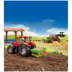 Red toy tractor with Amazone fertilizer broadcaster on dirt field, green hills, blue sky, and hay bales in the distance.