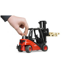 Red Bruder Linde Forklift toy with pallets, manipulated by a hand; realistic design, black and red color scheme.