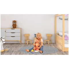 Heless potty with accessories beside a young girl holding a doll, surrounded by wooden furniture and a gray carpet.