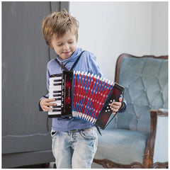 Young boy in blue shirt and jeans playing red and blue accordion with white buttons in front of a blue velvet chair.