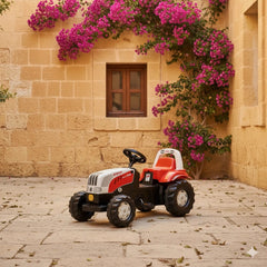 Rollykid Steyr 6165 Cvt tractor in red and white parked on stone tiles beside a plant with pink flowers.