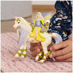 Fairy figurine in a yellow dress with wings rides a purple-maned unicorn on a soft pink surface.