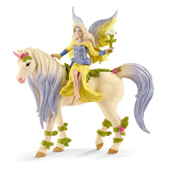 Fairy figurine with blonde hair in a yellow dress riding a white unicorn with a purple mane and green flower on its back.