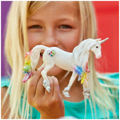 Rainbow Love Unicorn stallion figurine held by a smiling girl with blonde hair, wearing a light blue top.
