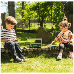 Theo Klein Bosch Work Box with two children holding a green container and scissors on a gray stone surface in a grassy area.