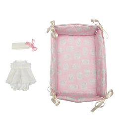 Pink textile cradle with decorative pattern, alongside a frilly white dress and matching headband with a pink bow.