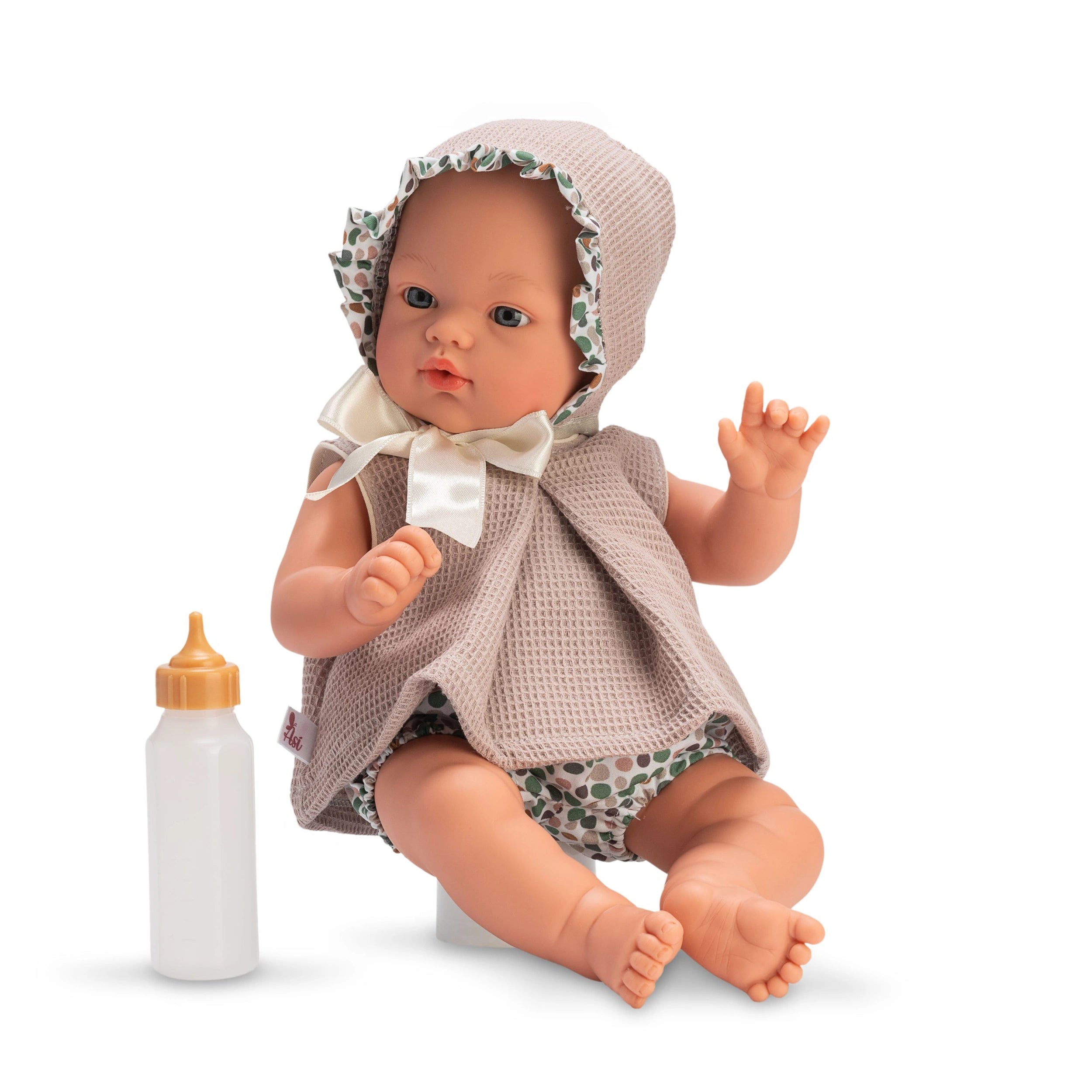 Koke from the Nude Dots Collection, wearing a bonnet and dress, with a feeding bottle beside him.