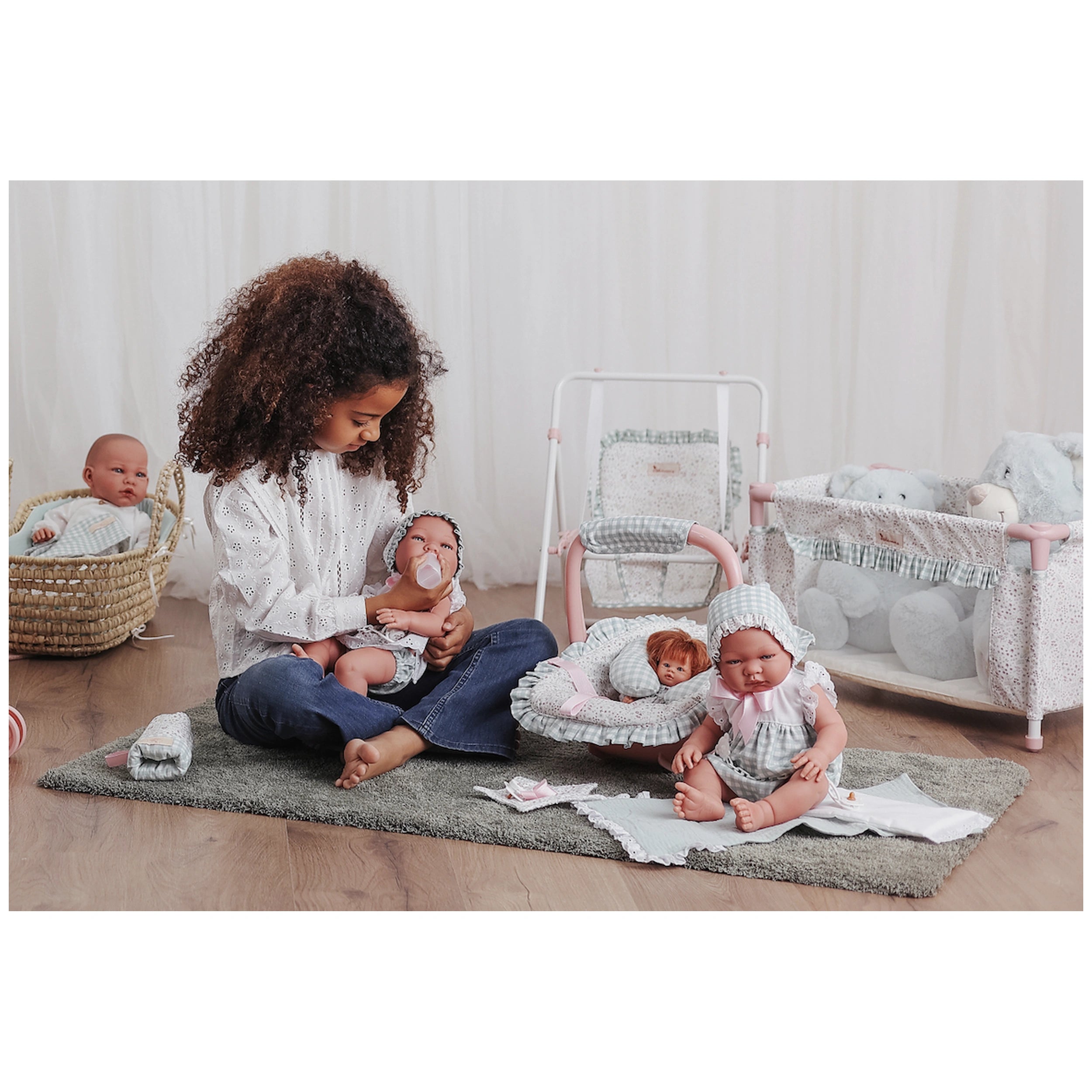 Young girl feeding a doll with a bottle, surrounded by several dolls and the ASI Dolls Maxi-Cosi with neck cushion.