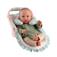 Doll in a pink Maxi-Cosi with a green neck cushion, wearing a green top and beige pants, against a floral-patterned ruffle.