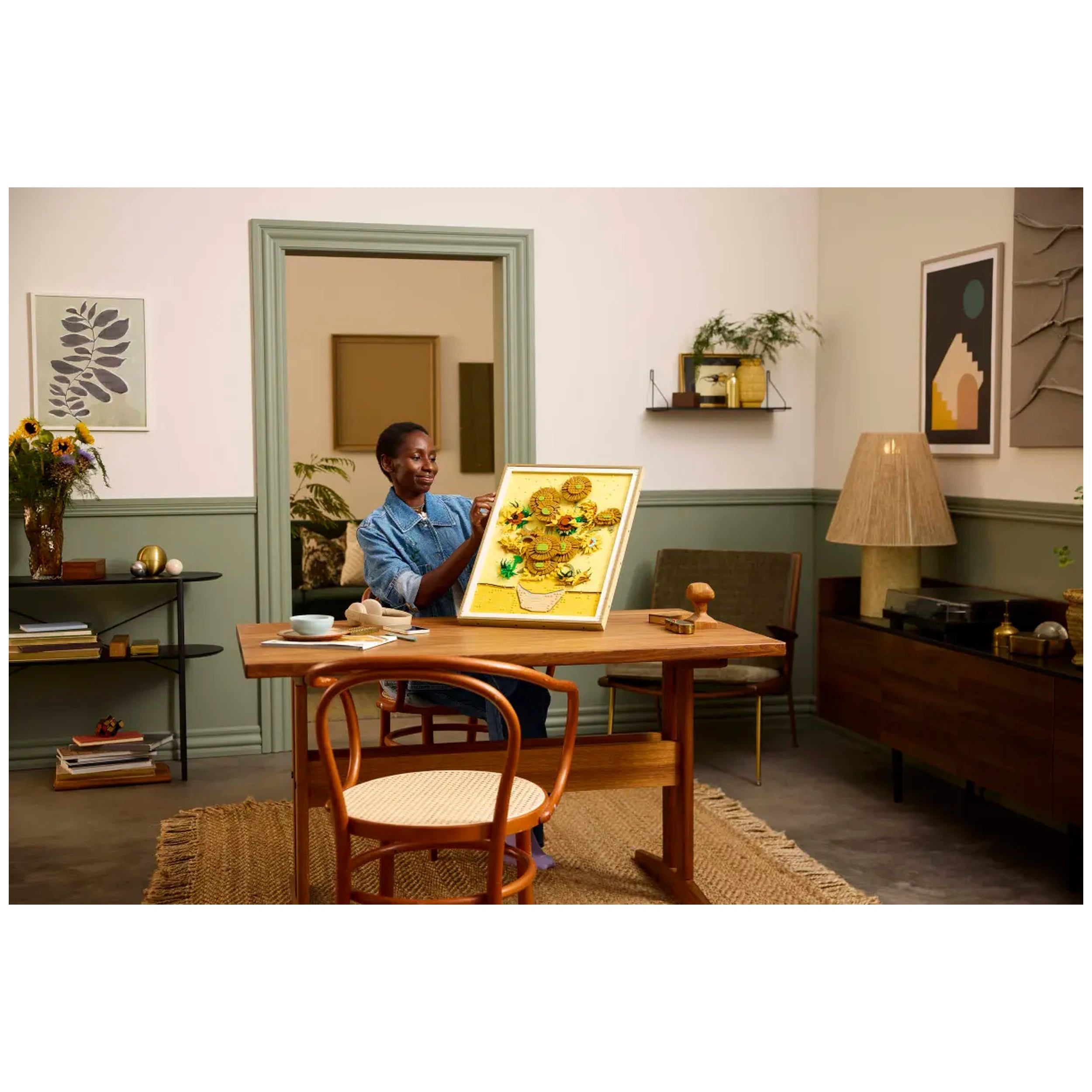 A person with short hair in a denim shirt admires a vibrant LEGO art piece of Van Gogh's "Sunflowers" in a cozy, modern living space with warm tones and natural light.