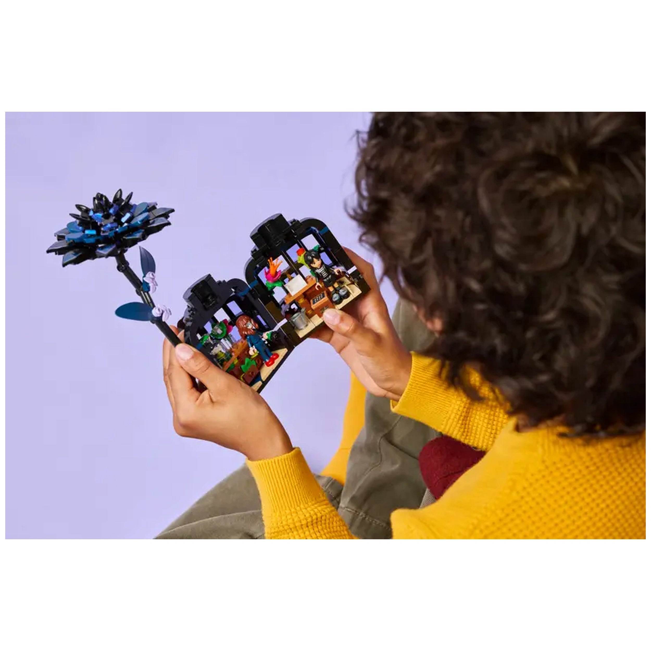 A child holds the LEGO Black Dahlia Flower set, showcasing a detailed black flower atop a mini conservatory with playful characters inside. The child wears a bright yellow sweater, highlighting their engagement in imaginative play against a soft purple background.