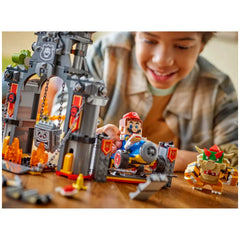 A child smiles while playing with the LEGO Bowser's Castle set, featuring Mario in a blue kart, a Thwomp, and Bowser, surrounded by flames and intricate structures.