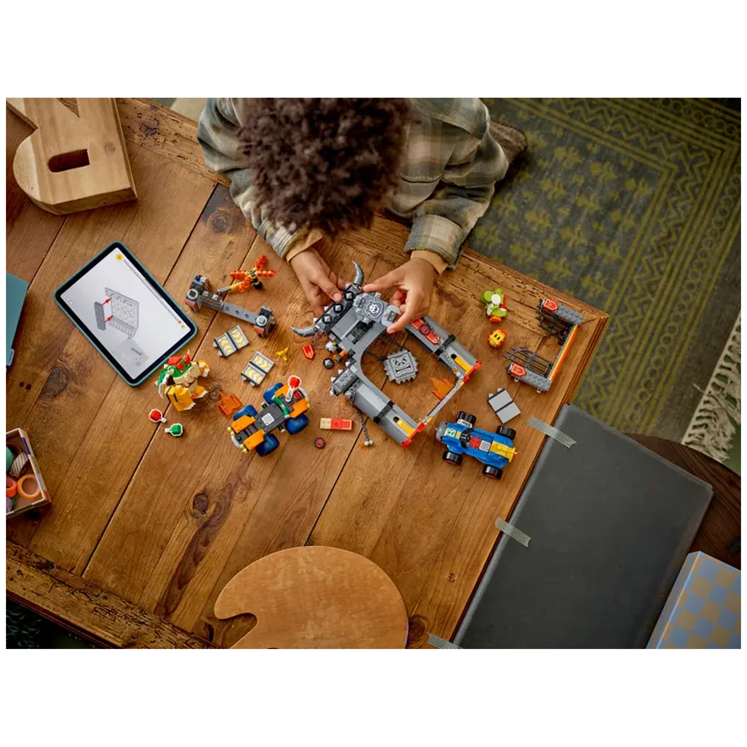 A child with curly hair is assembling LEGO pieces on a wooden table. Various colorful figures and parts are spread out, including cars and Bowser from the Mario universe. An illustrated instruction tablet is nearby, enhancing the play experience focused on Bowser's Castle.