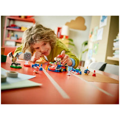 A child with curly hair smiles while playing with a LEGO Mario Kart set on a vibrant red table. The scene features an interactive Mario figure in a kart, alongside colorful track pieces, cones, and flags. Supplies like paper and tape are scattered nearby, adding to the creative environment.