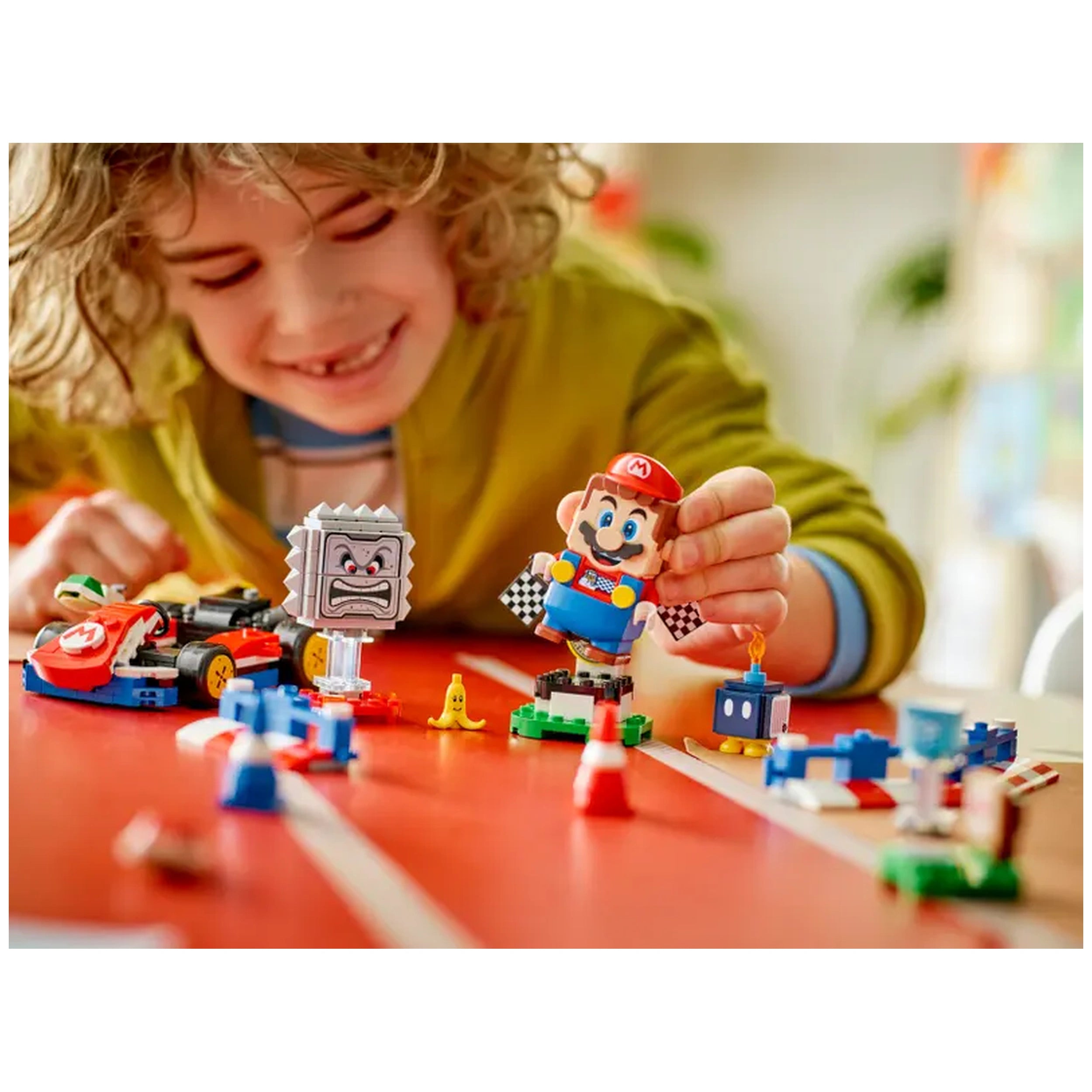 A young child with curly hair smiles while holding an interactive LEGO® Mario figure. The set features colorful blocks, a Bob-omb, a Thwomp, and a playful banana on a vibrant red surface. Nearby, a LEGO® car is positioned, part of a custom race track being built.