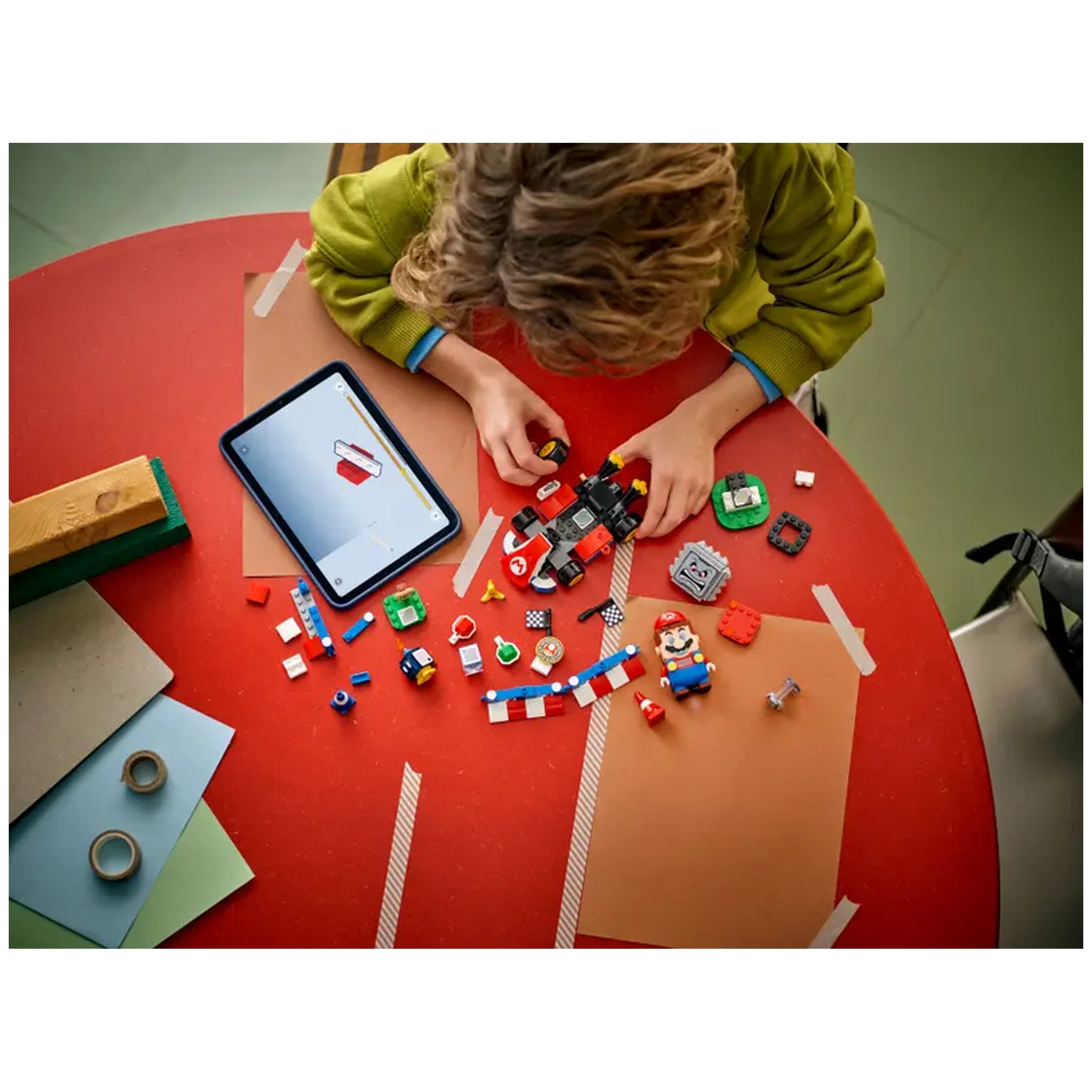 Child with curly hair assembling a LEGO Mario Kart playset on a red table, surrounded by colorful pieces and a tablet displaying instructions.