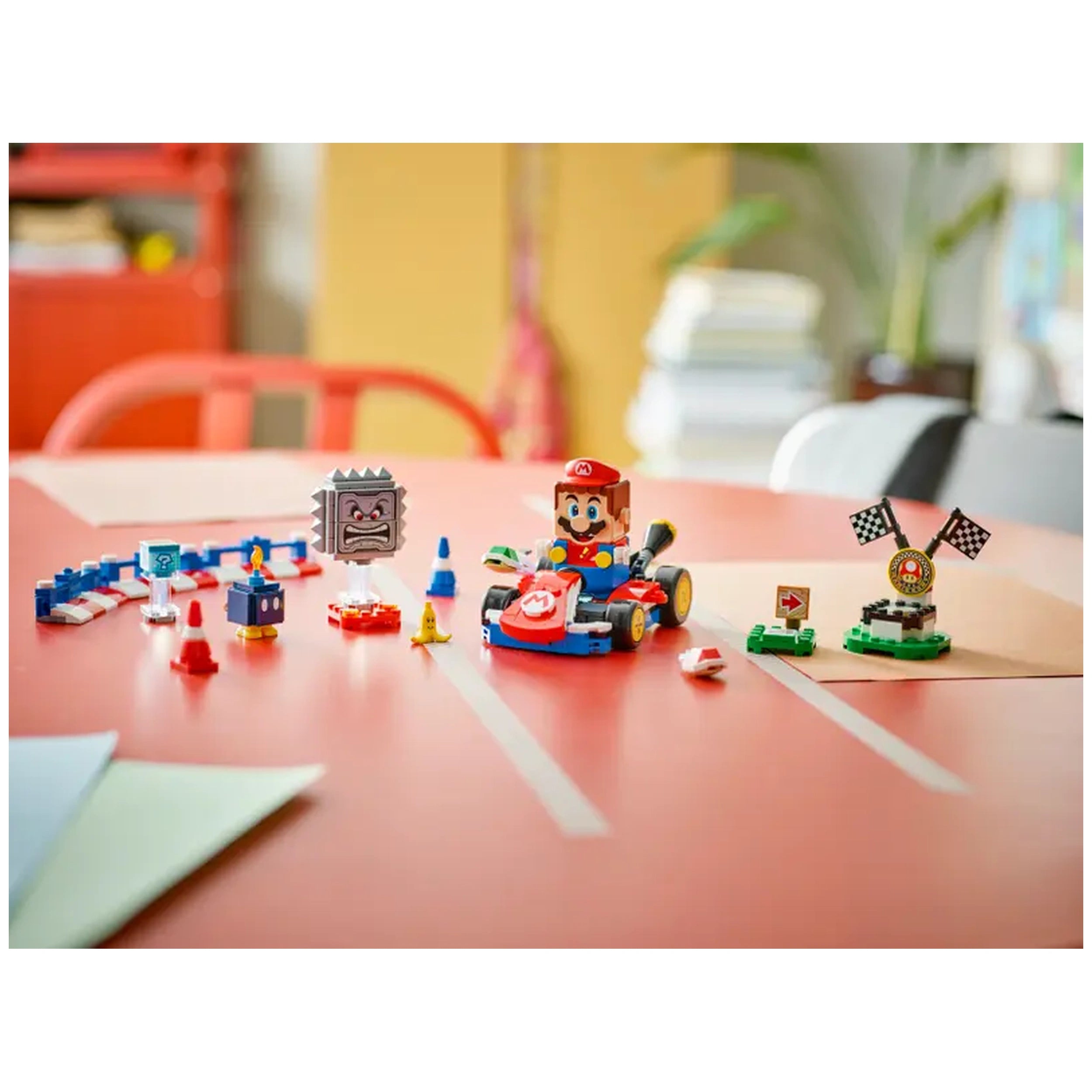 LEGO Mario in a red kart on a tabletop, surrounded by colorful obstacles, character figures, and racing flags, ready for play.