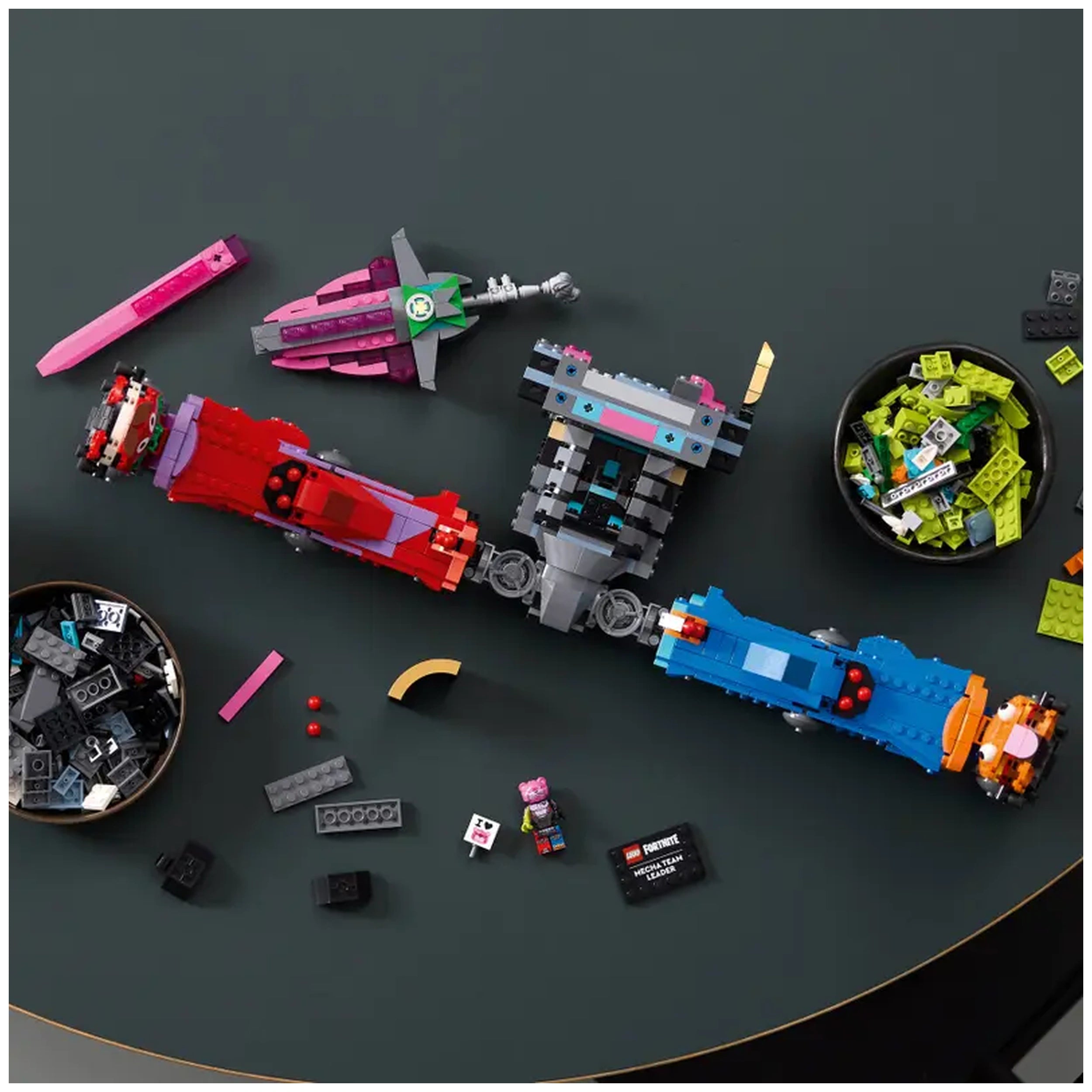 Colorful LEGO bricks arranged on a dark surface, featuring a large Fortnite Mecha Team Leader build in the center and various smaller pieces, including vehicles and accessories, alongside a bowl of green and black bricks. A Fortnite character figure is also present.