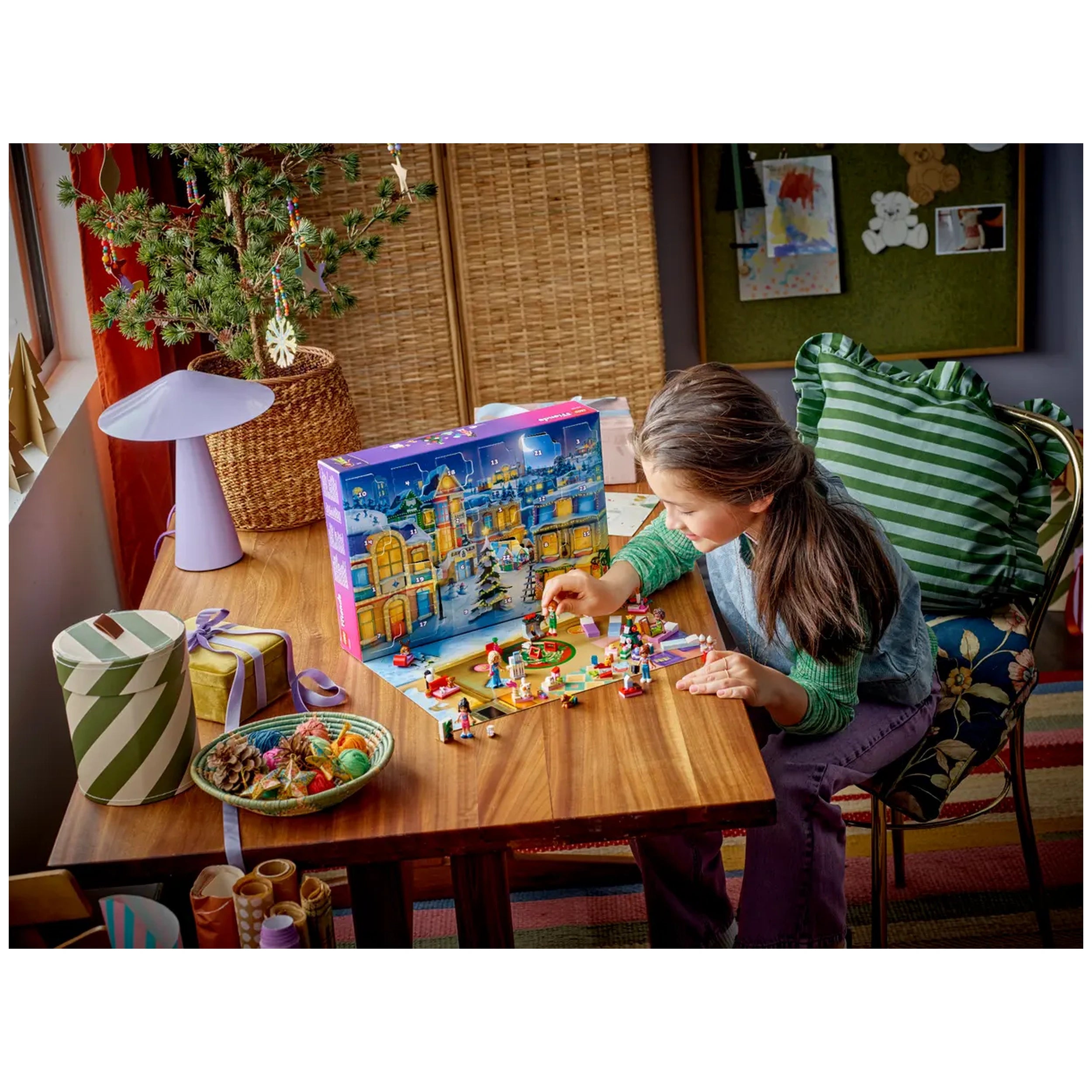 A girl with brown hair engages with the LEGO® Friends Advent Calendar on a wooden table, adding colorful pieces to a festive scene. Nearby, a small decorated tree and a patterned lamp create a cheerful atmosphere, while gift-wrapped boxes and a bowl of treats are visible.