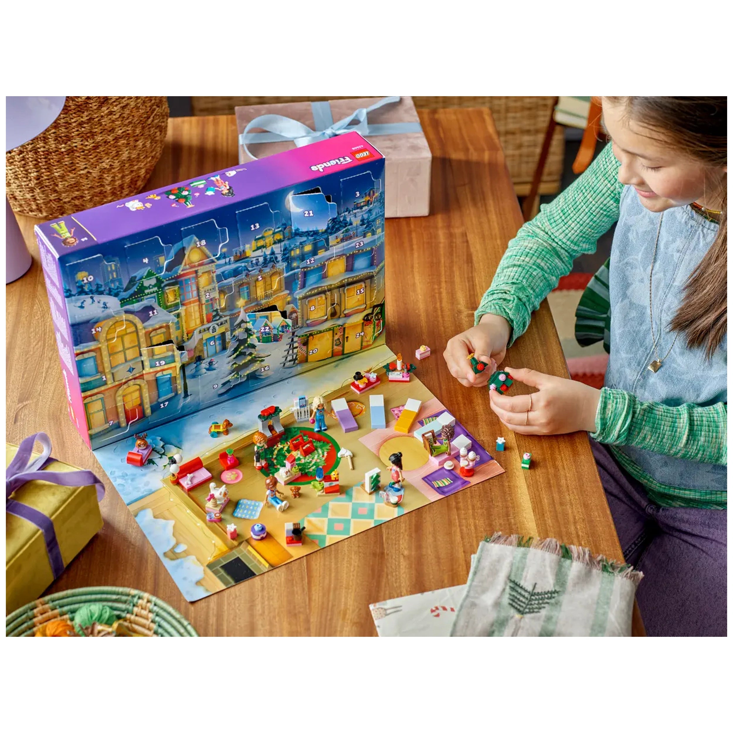 A child in a green sweater joyfully assembles the LEGO® Friends Advent Calendar 2025 on a wooden table. The calendar features a festive holiday scene with colorful mini-builds, including characters and animals, surrounded by presents and decor.