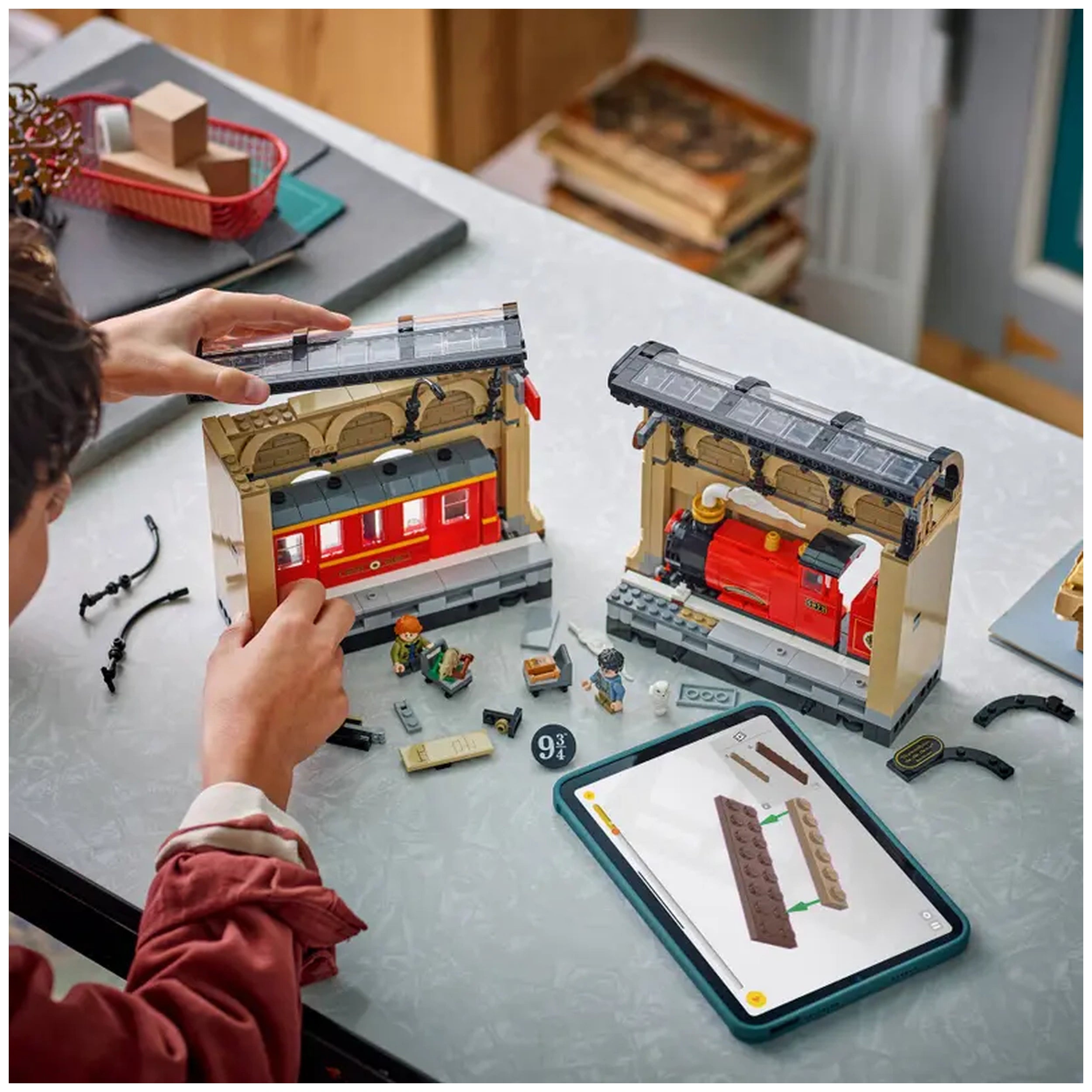 A child’s hand builds the LEGO Harry Potter Book Nook, featuring red train carriages and a locomotive against a light-colored table. Surrounding pieces and a tablet guide the assembly, with minifigures of Harry and Ron beside scattered LEGO bricks and accessories.