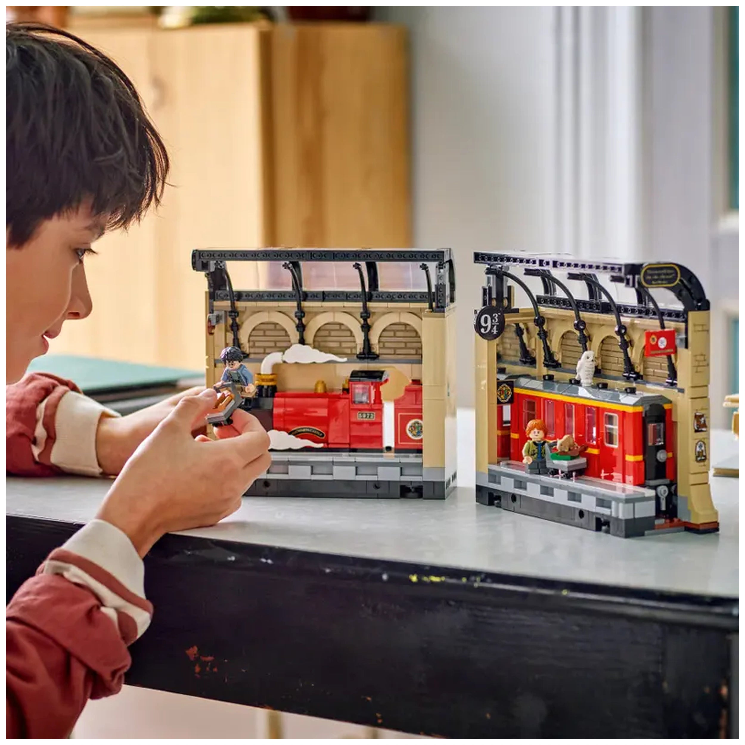 A child is focused on assembling a LEGO Harry Potter Book Nook, featuring a detailed Hogwarts Express scene with minifigures of Harry Potter, Ron Weasley, and their pets in a cozy indoor setting.