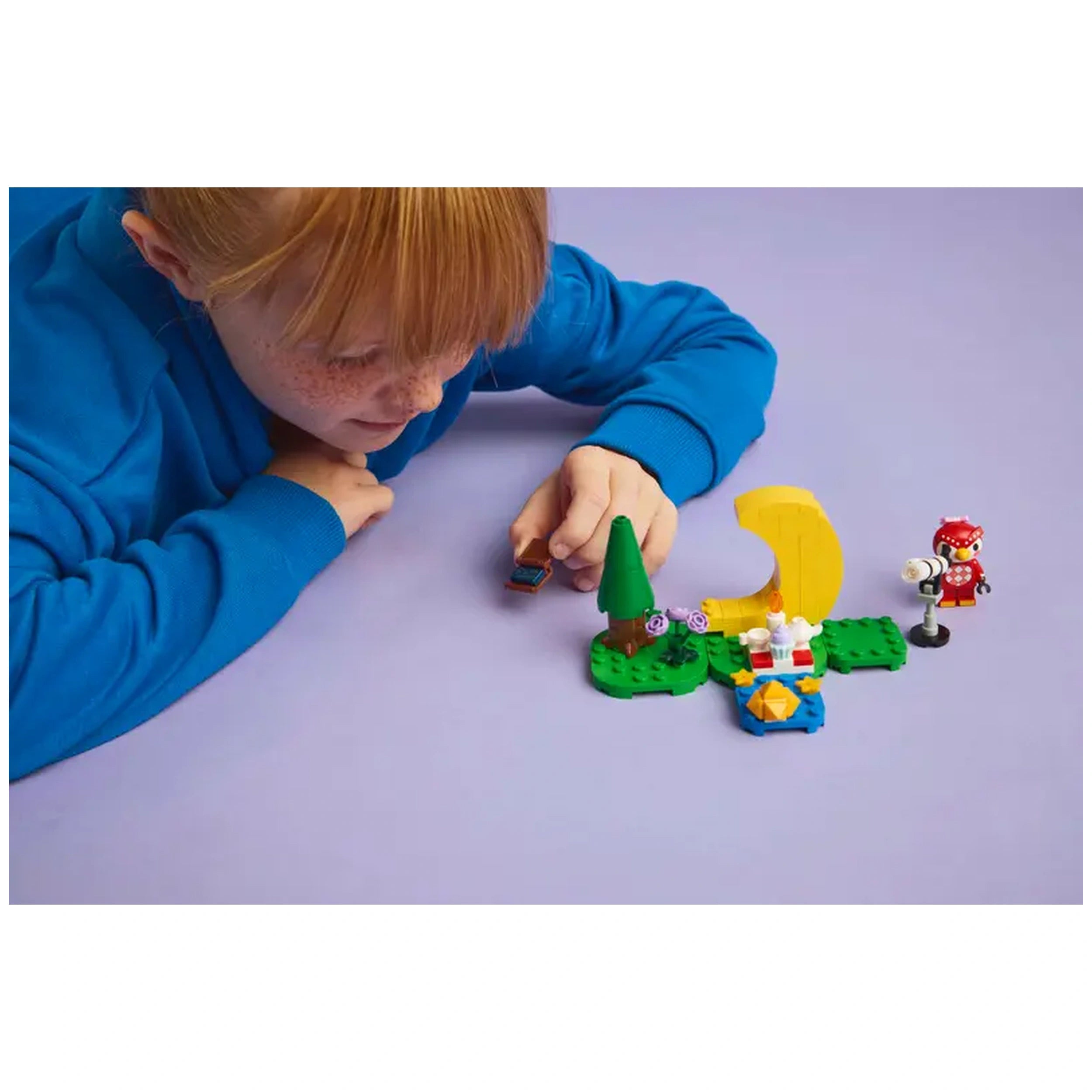 A young child in a blue sweatshirt leans over a colorful LEGO® set themed around Animal Crossing™, featuring a green base, a small tree, flowers, and characters, including a telescope and a crescent moon, on a light purple surface.