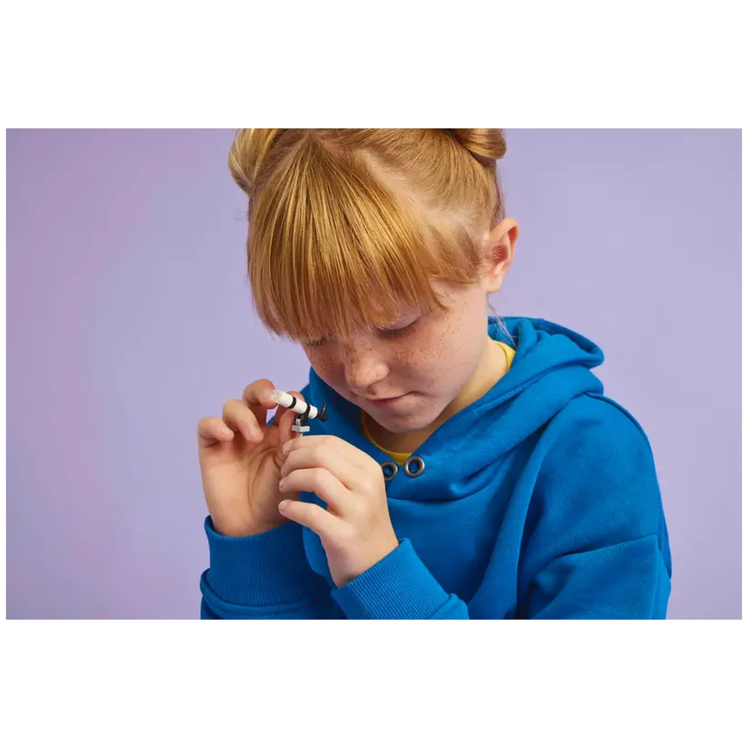 A child with red hair and freckles wearing a blue hoodie curiously holds a small toy telescope, focusing intently.
