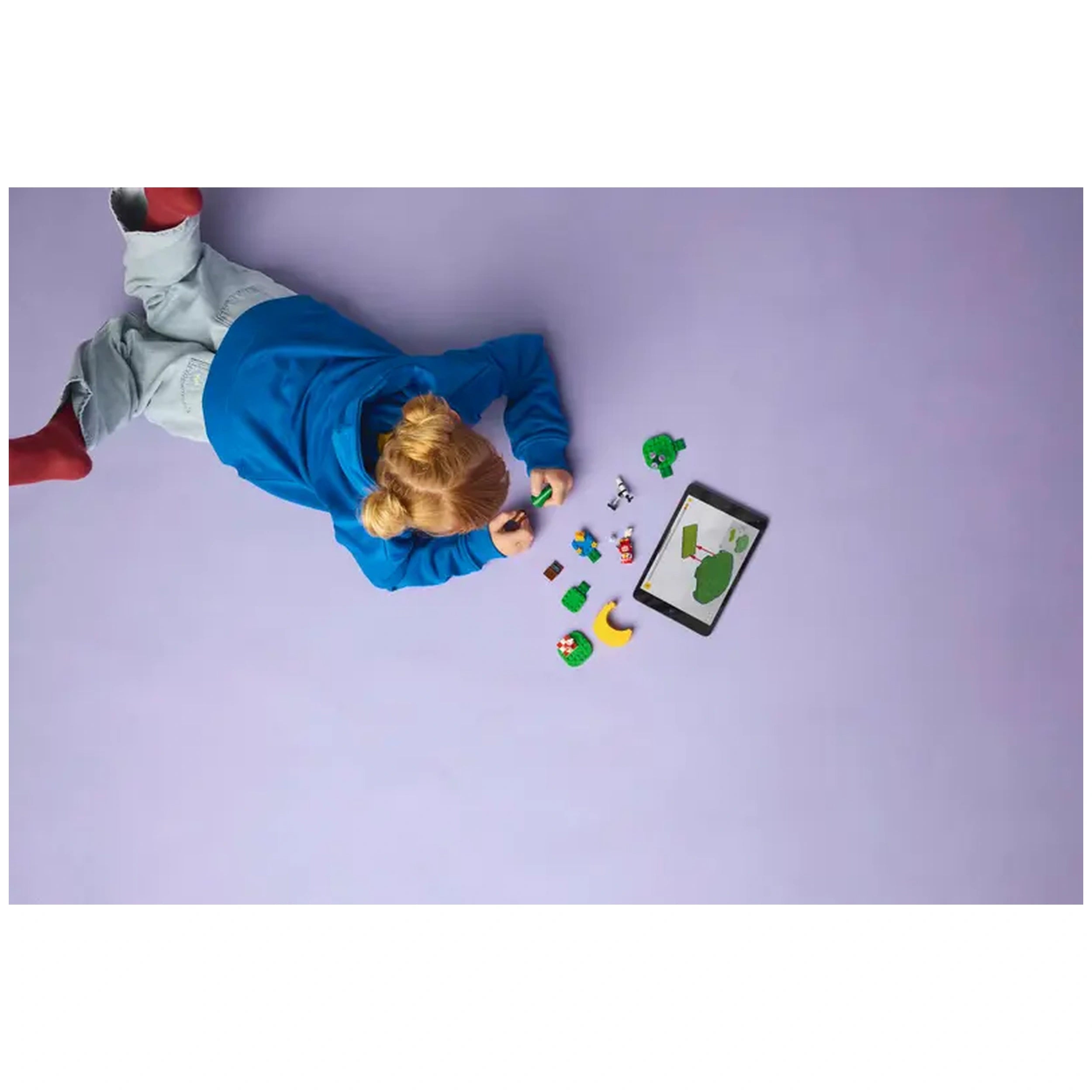 A child in a blue hoodie lies on a purple surface, focused on assembling colorful LEGO® pieces, with a tablet displaying a scene from Animal Crossing™ nearby.