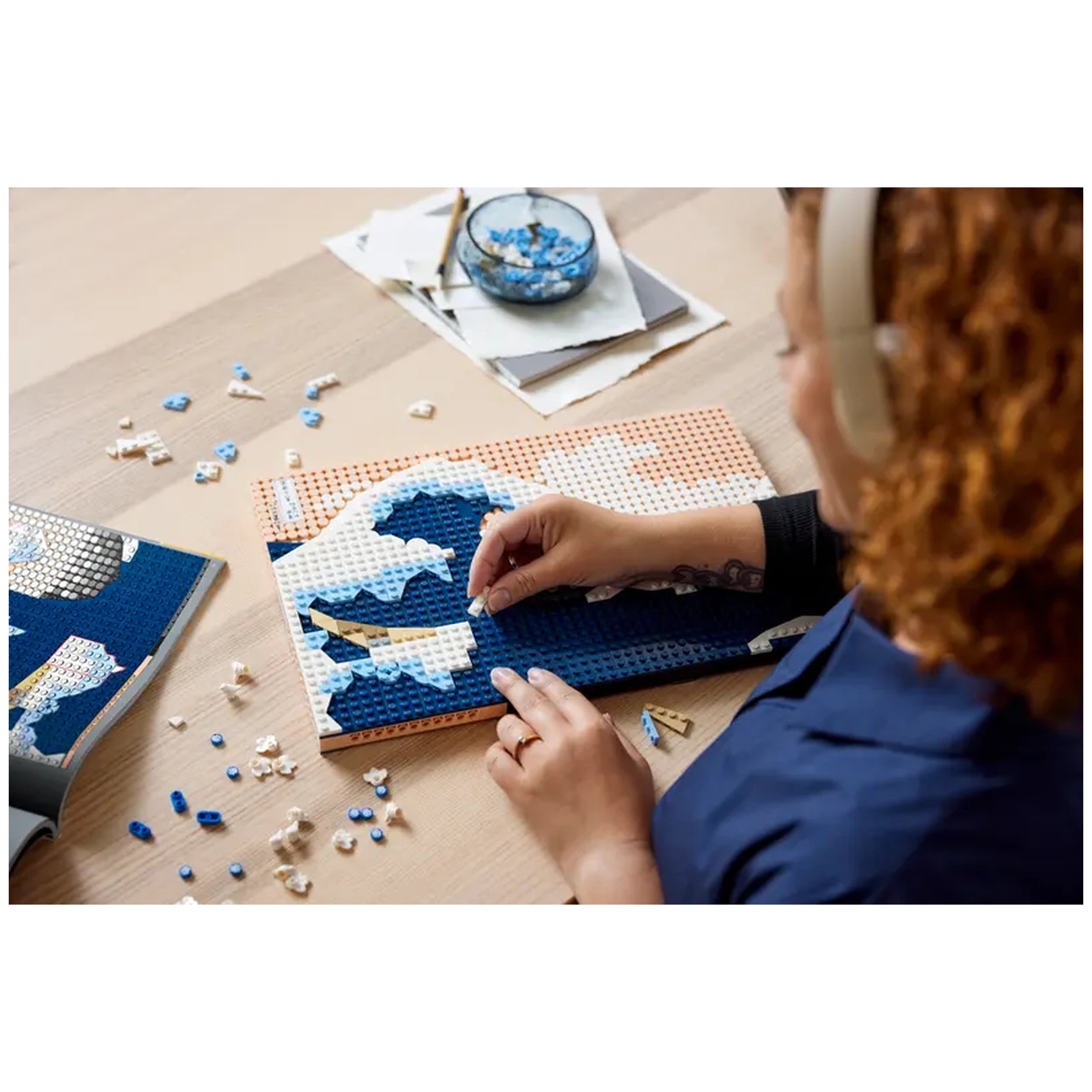 A person with curly hair sits at a wooden table, assembling a LEGO® Art set featuring Hokusai’s The Great Wave. Colorful pieces are scattered around, and they focus intently on placing blue and white bricks onto a base plate. A small bowl of blue pieces sits nearby, along with sketch papers and tools, creating an inviting workspace for creativity and mindfulness.
