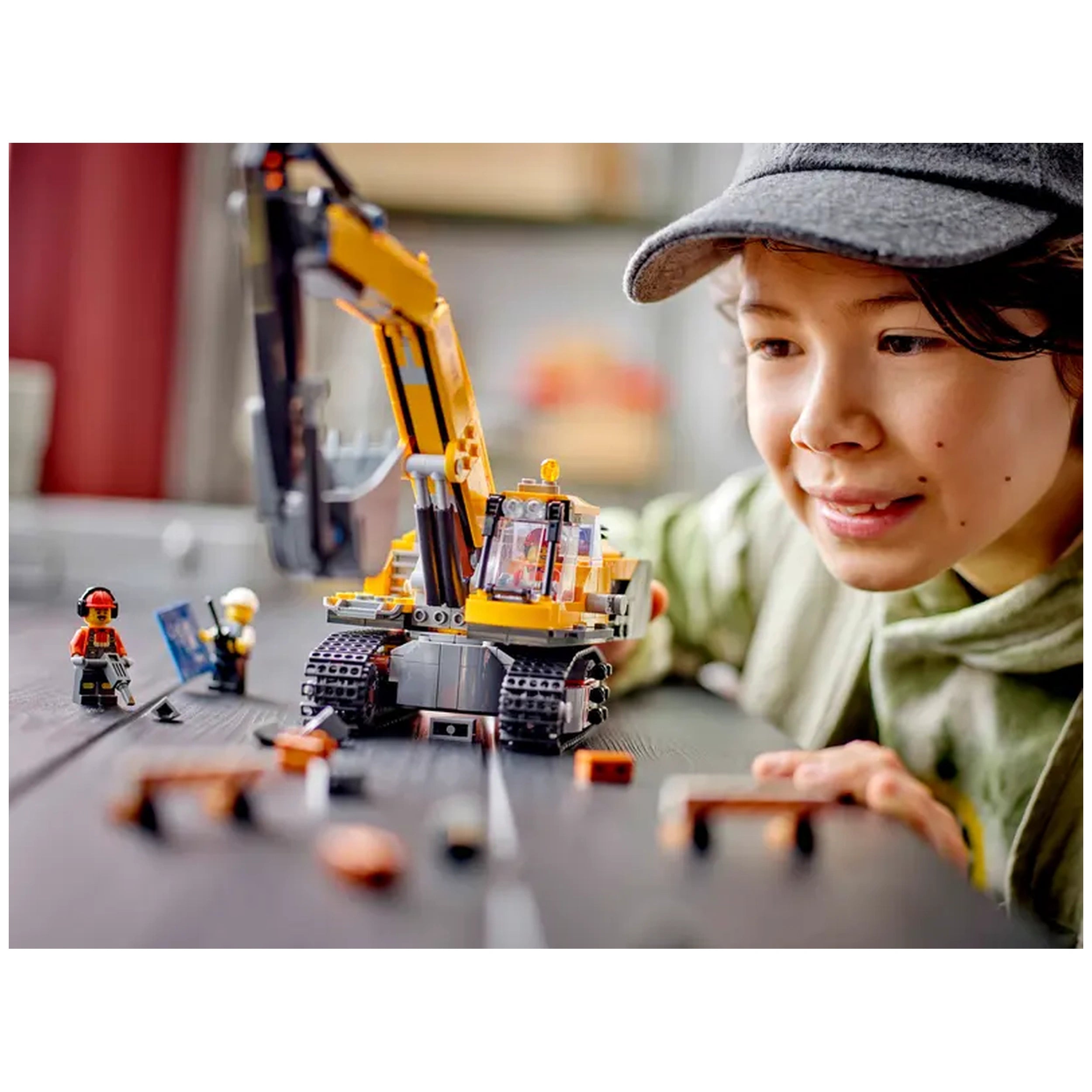 A child with a gray cap enthusiastically plays with a LEGO® City Yellow Construction Excavator, surrounded by minifigures and construction accessories on a wooden table.