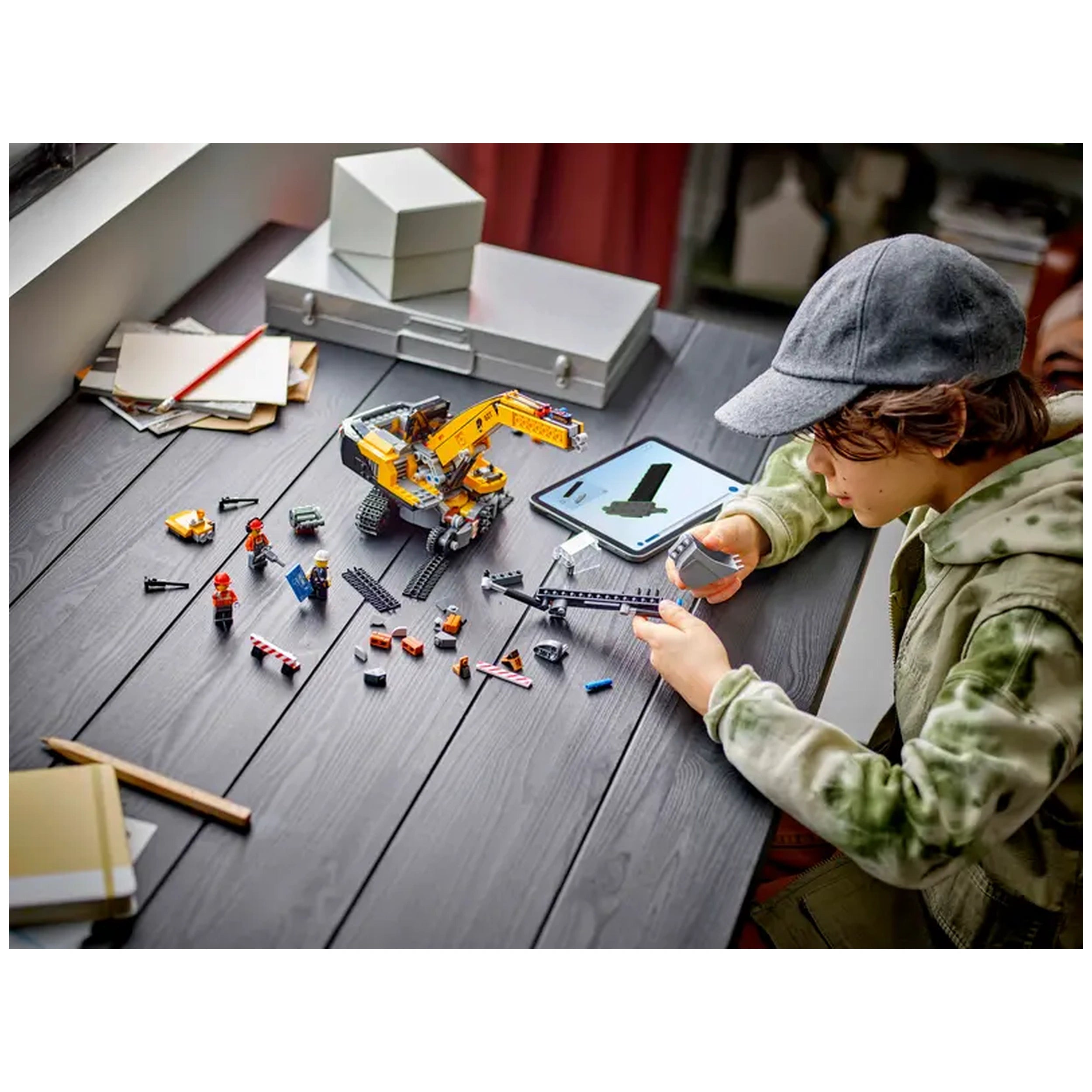 A child wearing a cap builds a LEGO® City Yellow Excavator on a dark wooden table. Surrounding them are various colorful blocks and accessories, including three minifigures and a tablet displaying building instructions.