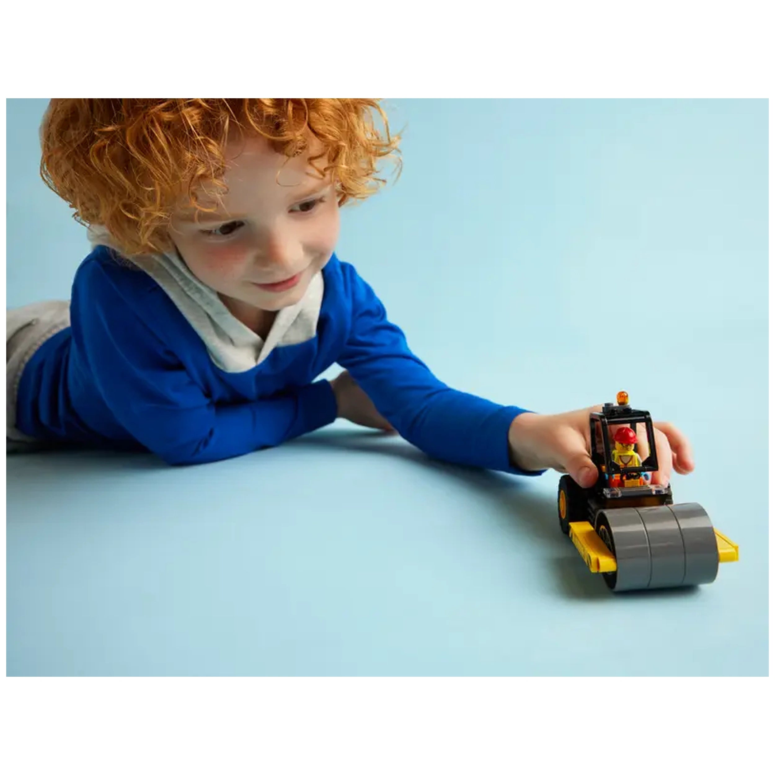 A young boy with curly red hair, wearing a blue sweatshirt, focuses on a LEGO® City Construction Steamroller toy with a construction worker minifigure.