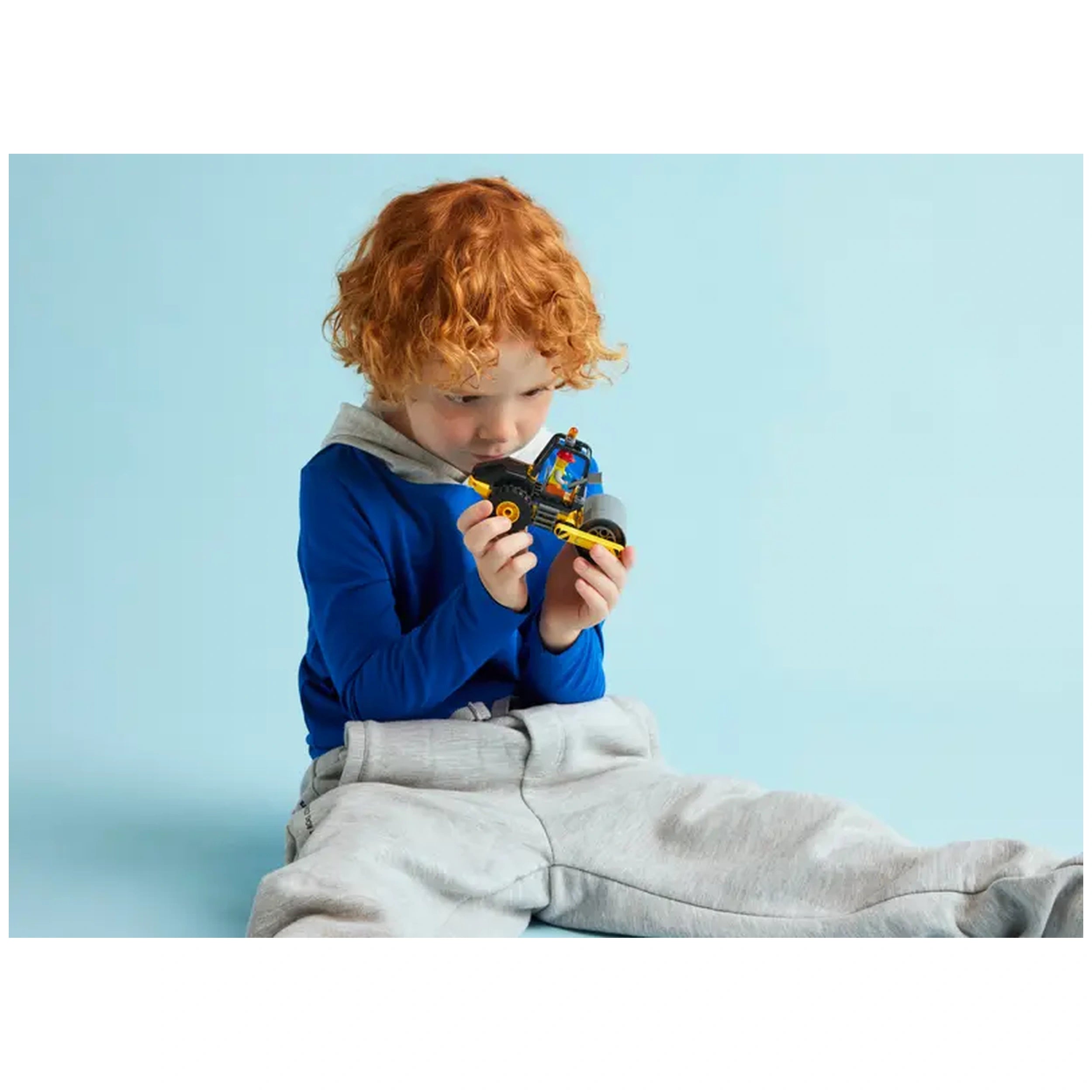 A young child with curly red hair sits on the floor, intently holding a LEGO City Construction Steamroller toy. The child wears a blue long-sleeve shirt with a gray hoodie and light gray sweatpants, against a soft blue background.