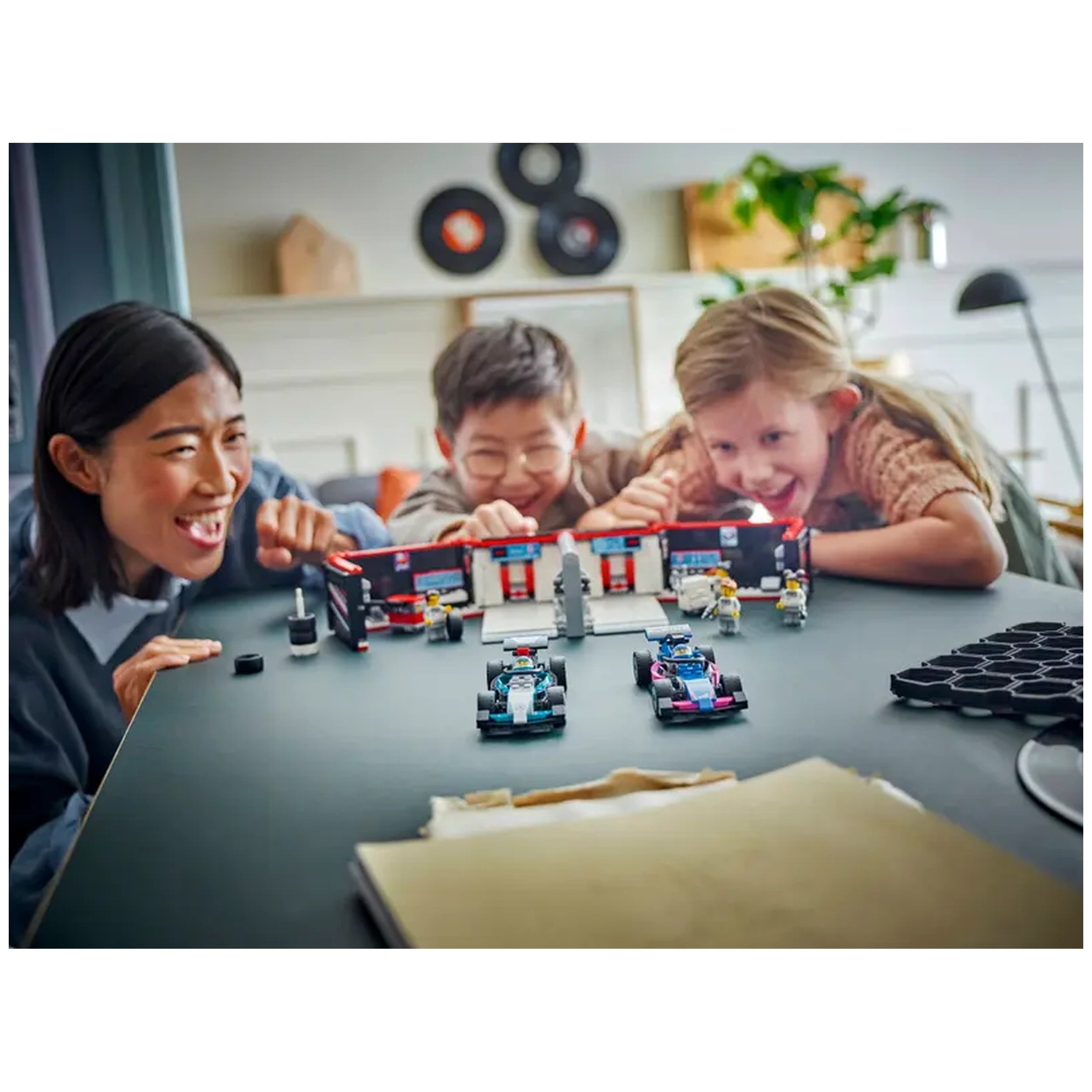 Three kids joyfully play with the LEGO® City F1® Garage set, featuring two race cars, a garage, and various mini-figures on a table.