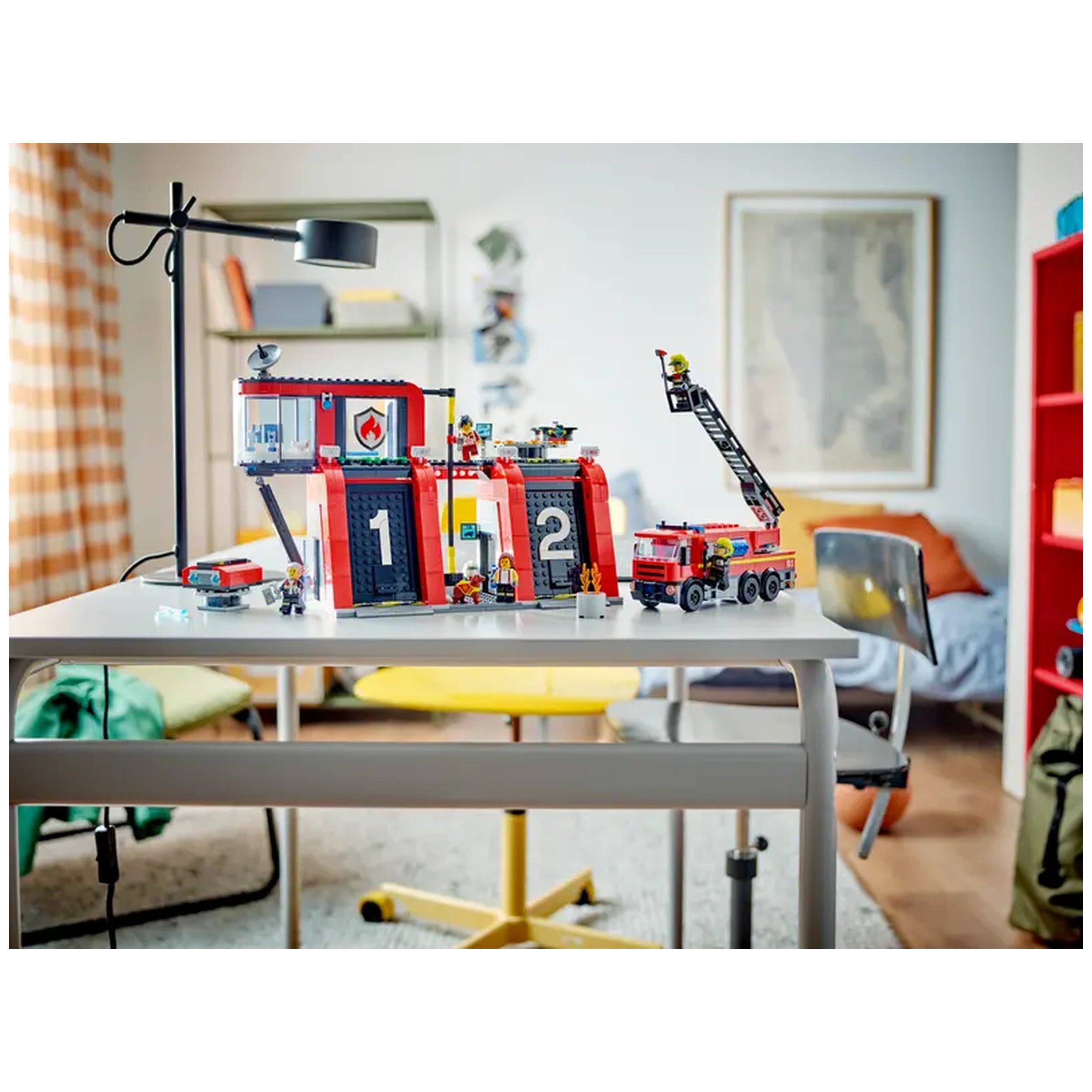 LEGO® City Fire Station set on a table, featuring a red building with numbered garage doors, a fire truck with a ladder, and five firefighter minifigures. A lamp and colorful furniture are visible in a cozy room.