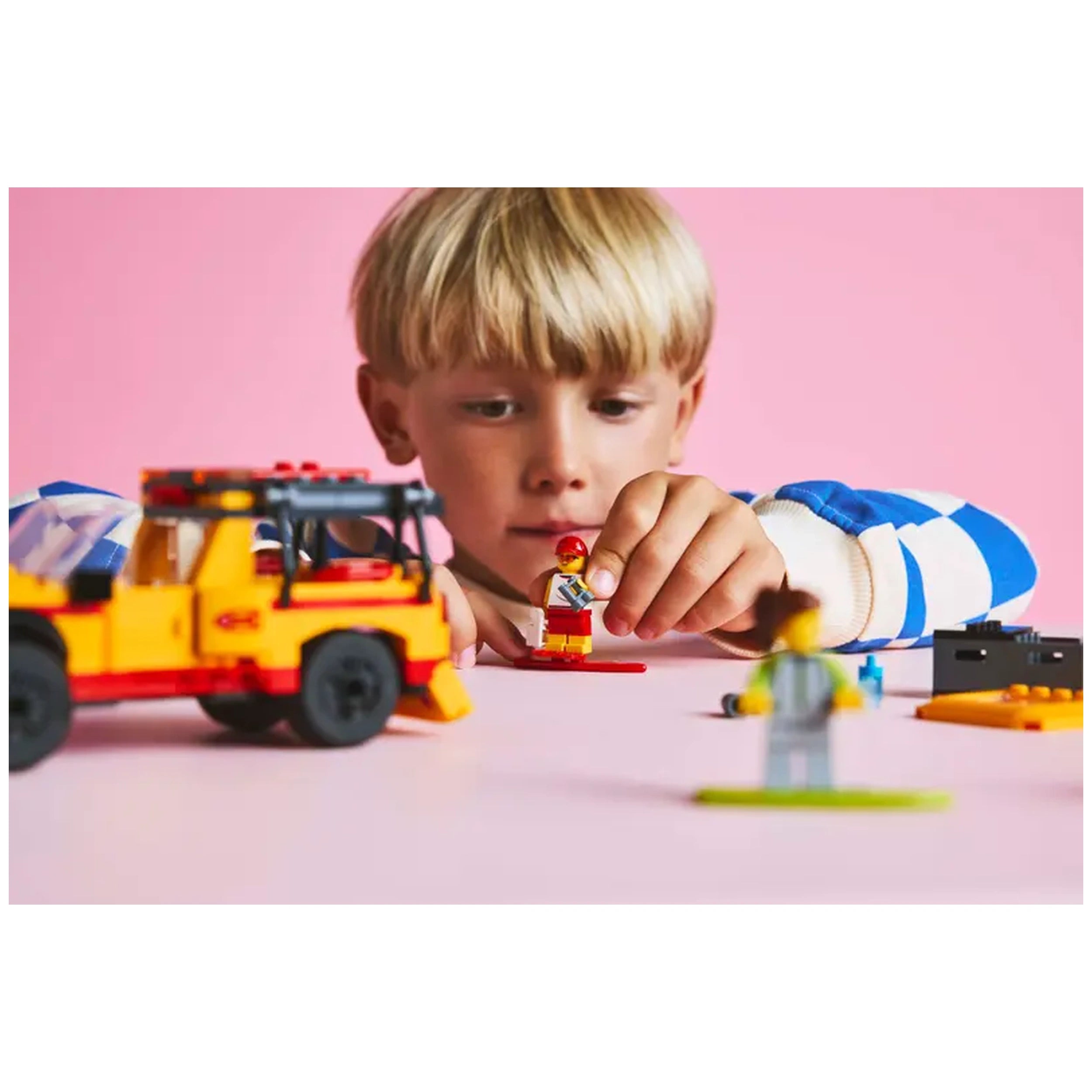 A young boy intently assembles a LEGO® City Lifeguard Beach Rescue Truck set, featuring vibrant yellow and red colors. He holds a lifeguard minifigure, surrounded by accessories like surfboards and a gray lifeguard figure on a pink surface.