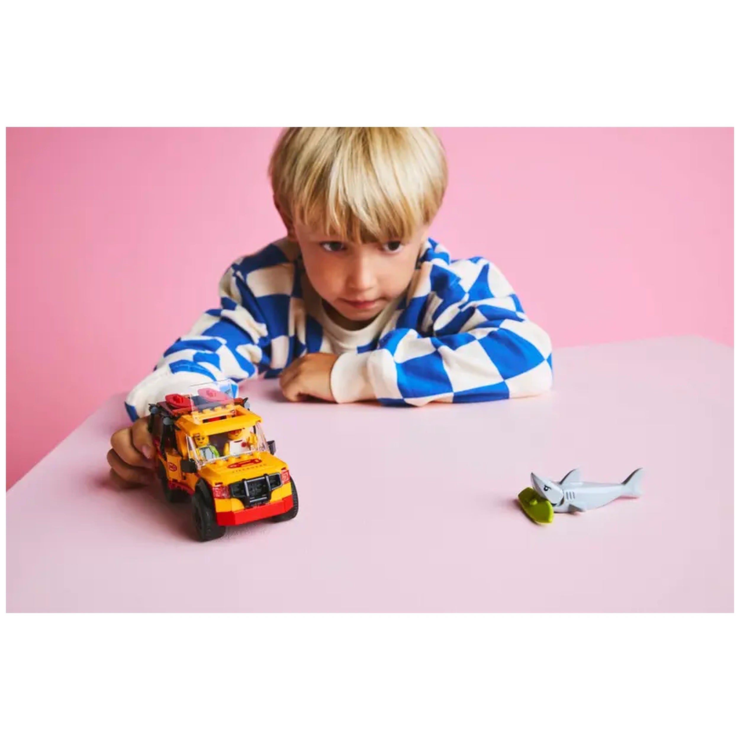 A child with blonde hair focuses on a vibrant LEGO® City Lifeguard Beach Rescue Truck, positioned on a pink table, alongside a gray shark figure.