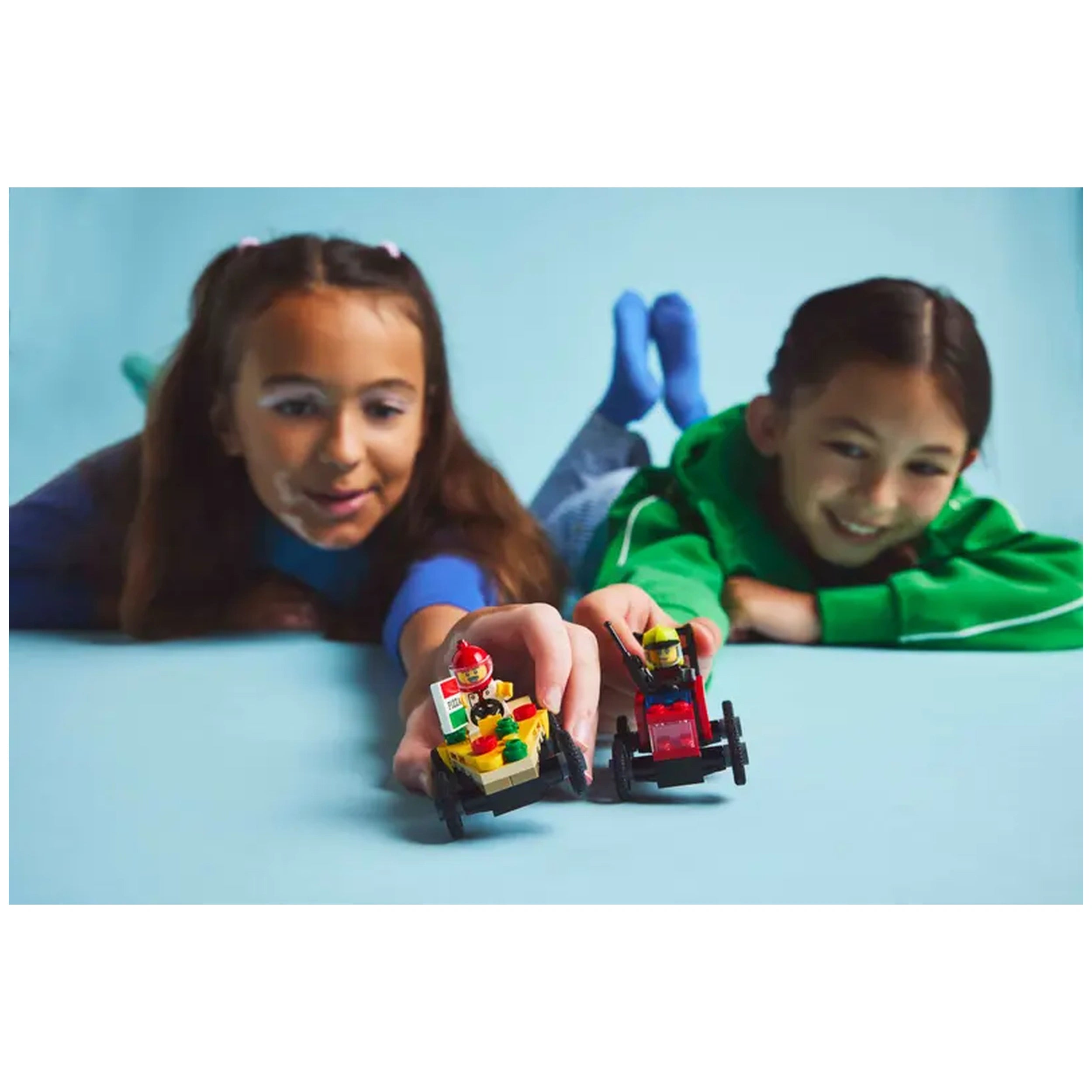 Two children excitedly reach for LEGO® race cars, one resembling a pizza slice and the other a fire truck, on a bright blue background.