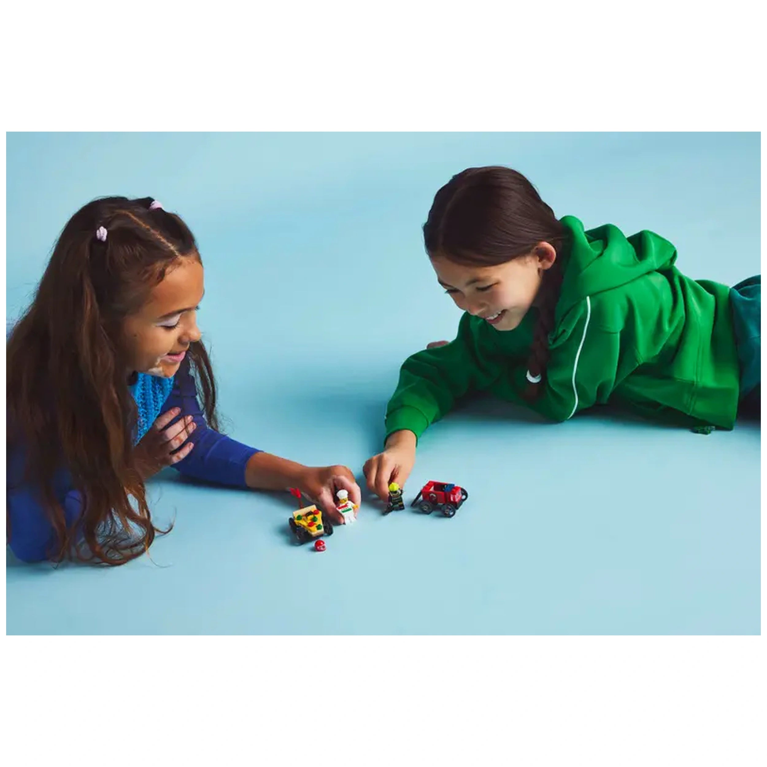 Two children play on a light blue surface, focused on LEGO® minifigures and race cars. One child, with long brown hair and a blue sweater, reaches towards the toys while smiling. The other child, wearing a green hoodie, leans closer, enjoying the interaction. The vibrant characters—a chef in a pizza car and a firefighter in a red truck—offer a glimpse of imaginative play and adventure.