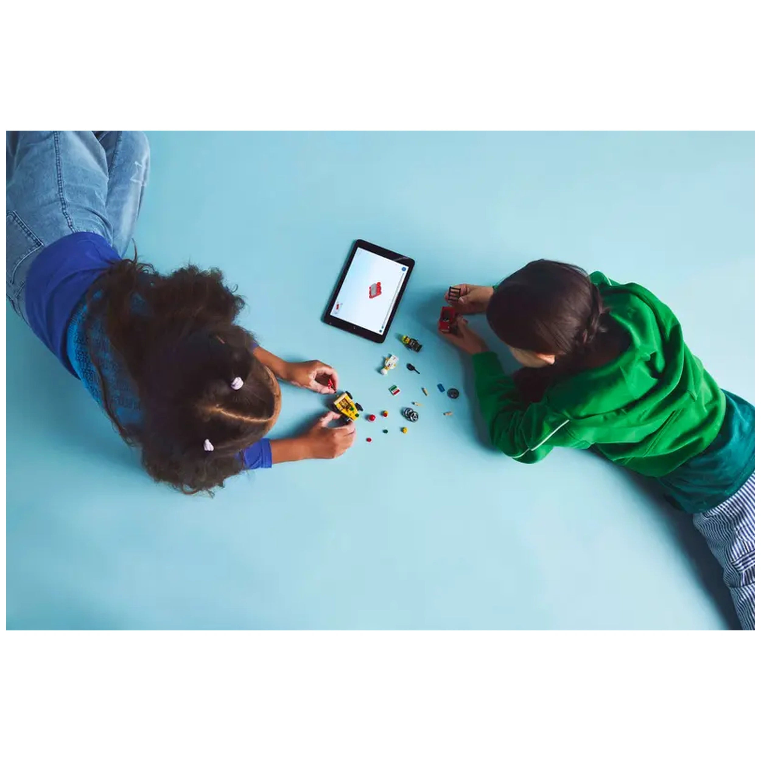Two children lying on a light blue surface, engaged in building LEGO® City vehicles. One child wears a blue shirt, holding a yellow car, while the other, in a green hoodie, assembles a red fire truck. An iPad displays LEGO® content nearby, surrounded by various colorful LEGO pieces.